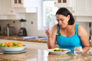 Mujer Sentada Al Mesón De La Cocina Mientras Come Un Plato De Ensalada Con Expresión De Disgusto. Medicamentos Como Ozempic Pueden Causar Incomodidad Al Comer Y Enfermedades Serias.